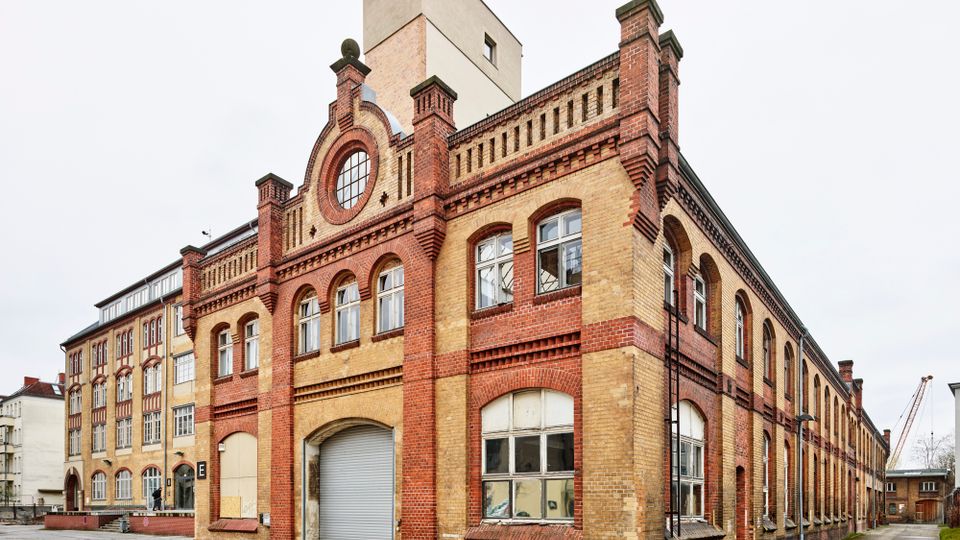 Backsteinbau eines ehemaligen Industriegebäudes mit roten und gelben Ziegelsteinen, Rundbogenfenstern und verziertem Giebel. Das Gebäude steht auf einem offenen Hof mit betonierter Fläche, daneben weitere ältere Backsteinstrukturen.