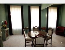 Ein Wohnzimmer mit grün gestrichenen Wänden, drei großen Fenstern mit weißen Vorhängen und dunklen Vorhangschals. Zentral steht ein runder Holztisch mit vier gepolsterten Stühlen, umliegende Möbel wie ein Kamin, ein Beistelltisch und ein roter Sessel ergänzen den Raum.