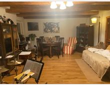 Wohnzimmer mit Holzboden und sichtbaren Holzbalken an der Decke. Der Raum ist mit einem Esstisch samt Stühlen, einer beige überzogenen Couch, einem Sessel mit gestreiftem Überwurf, Vitrinenmöbeln und Musikinstrumenten ausgestattet.