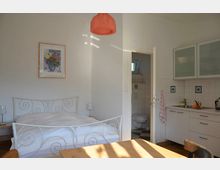 Helles Schlafzimmer kombiniert mit einer kleinen Küche, ausgestattet mit einem weißen Metallbett, einem Holztisch mit Stühlen, und einem weißen Küchenschrank mit Glastüren. Im Hintergrund führt eine Tür ins Badezimmer mit sichtbarer Toilette.