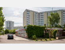 Straßenszene in einer Wohngegend mit mehreren Hochhäusern im Hintergrund, einem flachen Gebäude im Vordergrund mit der Aufschrift „Schul- und Sportjugendklub“, das teilweise von grünem Bewuchs bedeckt ist, sowie geparkten Autos entlang der Straße.