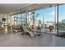 Wohnzimmer mit modernem Design, ausgestattet mit einem Liegestuhl auf einem Teppich, einer Lampe und Pflanzen. Große Glasfronten führen zu einem Balkon mit Blick auf Palmen und eine städtische Umgebung.