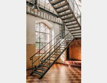 Industrieller Innenraum mit einer Metalltreppe, die diagonal nach oben führt, großen Fenstern mit Sprossen und einem Boden aus rot-gelbem Schachbrettmuster. Die Wände sind teils verputzt, teils mit Fliesen bedeckt, und eine große Hängelampe ragt von der Decke.