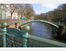 Grüne Eisenbrücke mit dem Schriftzug „Admiral-Brücke“ über einen ruhigen Stadtkanal, flankiert von Uferwegen, kahlen Bäumen und historischen Gebäuden im Hintergrund.