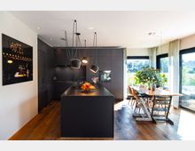 Moderne Wohnküche mit dunkler Einbauküche, einer schwarzen Kücheninsel und darüber hängenden Lampen, angrenzend an einen Essbereich mit Holztisch, Stühlen und großen Fenstern, die viel Tageslicht hereinlassen. Im Hintergrund ist eine grüne Pflanze auf dem Tisch und eine helle Landschaft durch die Fenster sichtbar.