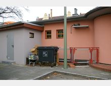 Außenbereich eines Gebäudes mit orangefarbener Fassade und kleinen Fenstern, daneben ein graues Nebengebäude mit Metalltür. Im Vordergrund steht ein Müllcontainer der BSR sowie ein Einkaufswagen mit Kartons und Gegenständen, umgeben von einem Gehweg und einem niedrigen Zaun.