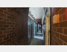 Schmaler Flur in einem Gebäude mit roten Klinkerwänden, grauen Metalltüren und mehreren Wandlampen an der Decke. Rechts ist ein orangefarbener Feuermelder angebracht, und der Boden ist aus Beton.