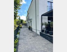 Eine gepflasterte Hofauffahrt neben einem modernen, weiß verputzten Gebäude mit Fenstern und einem Balkon. Links befindet sich ein Metallzaun mit angrenzender Pflanzenbepflanzung, zum Gebäude hin stehen Topfpflanzen und ein freistehender Sitzbereich mit Überdachung.