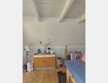 Innenraum eines Dachzimmers mit schräger weißer Decke und sichtbaren Deckenbalken, Holzfußboden und zweifarbig gestrichener Wand. Vor der Wand steht ein niedriges Holzsideboard mit Gegenständen und Lampe; rechts ist ein Bett mit blauem Bezug sowie mehrere abgestellte Taschen, Kartons und Stiefel zu sehen.