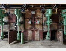 Industriehalle mit einer Ziegelwand, an der mehrere grüne, vertikale Maschinen mit Rohren, Ventilen und Schaltern befestigt sind. Unten sind offene Klappen und Metallstrukturen sichtbar, die zu einem alten mechanischen System gehören.
