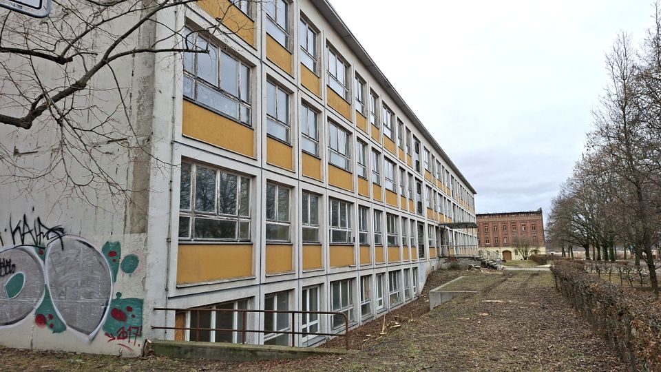 Langer, mehrstöckiger Gebäuderiegel mit Rasterfassade aus vielen Fenstern und gelb-weißen Fassadenelementen, an der Stirnseite mit Graffiti. Davor liegt ein schmaler Grünstreifen mit Weg und kahlen Bäumen; im Hintergrund ist ein weiteres Backsteingebäude zu sehen.