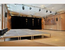 Heller Veranstaltungsraum mit Holzwänden und Bühne, die vor einem schwarzen Vorhang steht, dekoriert mit goldfarbenen Sternen. An der Decke sind Bühnenbeleuchtungen angebracht, an der rechten Wand befindet sich ein großes Holzrelief.