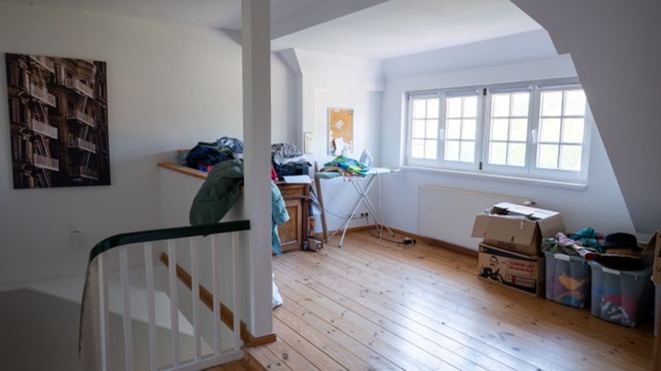 Dachgeschossraum mit schrägen Decken, Holzfußboden und großen, mehrteiligen Fenstern an der rechten Wand. Im Raum stehen ein Wäscheständer, Kisten und gelagerte Gegenstände; links befindet sich ein Treppengeländer und ein Wandbild.