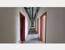 Ein langer, schlichter Flur mit weiß gestrichenen Wänden, rotem Türrahmen und mehreren offenen Türen. An der Decke verlaufen freiliegende Rohre und Kabel, während der Boden aus grauem Stein besteht.