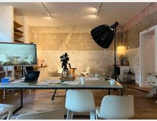 Ein modern eingerichteter Raum mit einer rustikalen Wand aus unverputztem Beton, ausgestattet mit einem großen Tisch, weißen Stühlen, einer Kamera auf einem Stativ und professionellem Studiolicht. Im Hintergrund befinden sich Regale, eine kleine Kommode und Dekoration wie eine Lampe und Blumen.