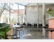 Helles, modernes Café mit großen Glasfenstern, die den Blick ins Freie freigeben. Innen befinden sich weiße Tische, schwarze Stühle, Betonwände und einzelne Pflanzen.