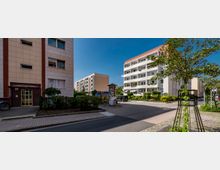 Wohnsiedlung mit mehreren mehrstöckigen Gebäuden aus heller Fassade, bepflanzten Grünflächen und einem zentralen gepflasterten Weg. Im Vordergrund sind kleine Bäume und eine Litfaßsäule sichtbar, umgeben von gepflegter Vegetation und einem Zugang von der Hauptstraße.