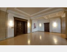 Innenbereich eines öffentlichen Gebäudes mit einem großen, hellen Raum. Zu sehen sind zwei dunkle Holztüren, eine davon mit der Aufschrift „Magistratssitzungszimmer“, helle Wände mit Wandlampen und schlichte Stuckverzierungen an den Deckenrändern.