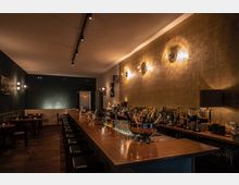 Eine stilvolle Bar mit langer Theke aus Holz, flankiert von schwarzen Barhockern. Im Hintergrund sind dunkle Wände, kleine Tische mit Stühlen sowie dekorative Beleuchtung und eine Wand mit goldener Struktur zu sehen.