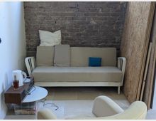 Ein kleiner Raum mit einer freiliegenden Backsteinwand, in dem ein beiges Sofa mit Kissen steht. Links daneben befindet sich ein Holzbeistelltisch mit einem Wasserkocher und einem Glas Tee; daneben liegt ein Sessel im Vordergrund. Auf der rechten Seite sind Holzplatten an der Wand angelehnt.