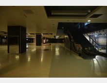 Ein leerer, weitläufiger Innenraum eines Einkaufszentrums oder Geschäftsgebäudes mit hellen Laminatböden und dunklen Säulen. Rechts im Bild befindet sich eine ungenutzte Rolltreppe, daneben Glasfenster mit Blick auf eine beleuchtete Außenszene.