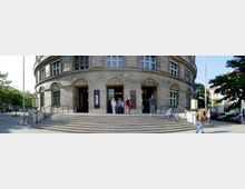 Halbrundes Gebäude mit Natursteinfassade, großen Fenstern und dekorativen Bögen über den Eingängen. Eine breite Treppe mit Handläufen führt zu den Eingängen, während Menschen davor stehen oder vorbeigehen; Umgebung mit Bäumen und parkenden Autos.