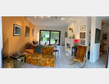 Wohnzimmer mit buntem Sofa, dekoriert mit floralen Mustern, und einem Sessel vor einer weißen Wand mit Kamin und Bildern. Im Hintergrund sind eine große Glas-Schiebetür mit Blick auf einen Garten sowie helle Holzböden und eine Treppe sichtbar.