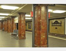 Untergrundbahnhof mit Säulen aus braunen, verzierten Kacheln, hellem Deckenlicht und Sitzgelegenheiten aus Metall. Im Hintergrund befindet sich ein Schild mit der Aufschrift „Fehrbelliner Platz“.
