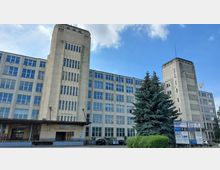 Großes, mehrstöckiges Industriegebäude mit rechteckigen Fenstern und zwei markanten Türmen, die vertikale Fensterreihen aufweisen. Im Vordergrund befinden sich einige Bäume, geparkte Autos und Hinweisschilder mit Firmennamen.