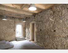 Ein rustikaler Innenraum mit groben Natursteinwänden, einem hölzernen Balkendecken und einem Boden aus Ziegelsteinen. Im Raum befinden sich ein großes Fenster mit einer Treppe darunter sowie eine Türöffnung ohne Tür.