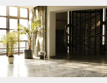 Heller Innenraum mit Marmorboden, großen Fenstern, die viel Tageslicht hereinlassen, zwei Topfpflanzen und einem modernen Treppenhaus mit Metallgeländer im Hintergrund.