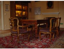 Ein klassisch eingerichteter Raum mit einem dunklen Holztisch und passenden Stühlen mit gestreiften Polstern, umgeben von einem roten, gemusterten Teppich. Im Hintergrund steht ein antiker Bücherschrank mit beleuchteten Bücherregalen sowie gerahmte Bilder und ein Kamin an der beigen Wand.