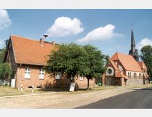 Backsteingebäude mit Satteldach neben einer Kirche mit Spitzdach und Turm aus Backstein und Feldsteinen; im Vordergrund eine asphaltierte Straße und ein Baum. Der Himmel ist leicht bewölkt.