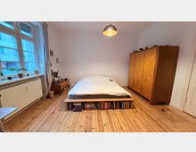 Schlafzimmer mit Holzboden, einem zentral positionierten Bett mit integriertem Bücherregal am Fußende, einem großen Holzkleiderschrank rechts und einem Fenster mit Pflanzen auf der Fensterbank links. Eine hängende Deckenlampe beleuchtet den Raum.