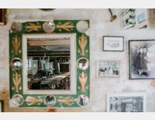 Ein grüner Wandspiegel mit goldenen Verzierungen reflektiert einen Raum mit einer langen Tafel und vielen Stühlen in rustikaler Umgebung. Die Wand um den Spiegel ist mit verschiedenen gerahmten Bildern dekoriert.