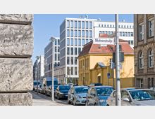 Straßenszene in einer europäischen Stadt mit moderner und klassischer Architektur: Im Vordergrund parken Autos entlang einer gepflasterten Straße, rechts ein gelbes historisches Gebäude mit rotem Dach, dahinter mehrstöckige weiße Bürogebäude mit Glasfassaden.