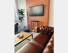 Wohnzimmer mit Backsteinwand, an der ein Flachbildfernseher hängt. Im Vordergrund ein braunes Ledersofa, ein Holztisch mit Metallrahmen, und ein Regal mit Kaminholz; seitlich eine große Zimmerpflanze in einem weißen Topf.