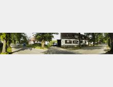 Straßenszene in einem Dorf mit Kopfsteinpflasterstraßen, gesäumt von Bäumen. Rechts sind Häuser mit Satteldächern und weißen Fassaden zu sehen, links steht ein rosa Haus in einem begrünten Bereich, und mehrere parkende Fahrzeuge sind sichtbar.