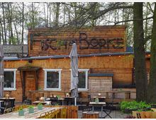 Holzgebäude mit der Aufschrift „Fischer Borke“, umgeben von Bäumen. Davor befindet sich ein Außenbereich mit Tischen, Stühlen, Sonnenschirmen und kleinen Pflanzen.