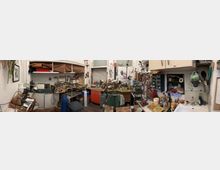 Überfüllte Werkstatt mit mehreren Arbeitstischen, Regalen und Schubladenschränken, bedeckt mit Werkzeugen, Kabeln und Kleinteilen. Im Raum sind zudem ein Waschbecken mit Küchenutensilien, Hängeschränke, Pinnwände sowie Fenster und eine Hochlagerfläche mit Stoffen und Kartons zu sehen.