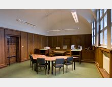 Ein Raum mit Holzwänden und einem gewölbten weißen Deckenbereich, ausgestattet mit einem Konferenztisch und mehreren Stühlen in der Mitte. Auf der rechten Seite befinden sich große Fenster, die Tageslicht einlassen, und im Hintergrund stehen mehrere Schreibtische mit Bürostühlen und Arbeitsmaterialien.