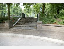 Treppenaufgang in einem Park mit mehreren Stufen, flankiert von niedrigen Backsteinmauern und grünem Metallgeländer. Im Hintergrund sind ein umzäunter Bereich sowie Bäume und Sträucher zu sehen.