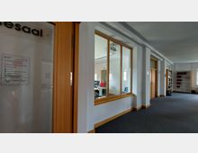 Flur in einem Verwaltungsgebäude mit Teppichboden, weißen Wänden und Holztüren. Links befinden sich Büros mit großen Glasfenstern; rechts sind Bücherregale und Fenster sichtbar.