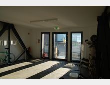 Ein heller Innenraum mit Betonboden, großen Glastüren, die zu einem Balkon führen, und sichtbaren Stahlträgern an den Wänden. Im Raum befinden sich ein grünes Fahrrad, ein Stuhl, ein Regal mit Büchern und Feuerlöscher an der Wand.