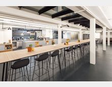 Großer, moderner Gemeinschaftsraum mit einer langen Tischreihe und schwarzen Barhockern, dekoriert mit kleinen Pflanzen und Keramikgegenständen. Im Hintergrund eine große offene Küche mit kaffeebarartigen Elementen, Regalen und Kaffeemaschinen.