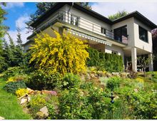 Modernes Wohnhaus mit zwei Etagen und Flachdach, umgeben von einem dicht bepflanzten Garten mit gelb blühendem Strauch, Rasenflächen und anderen Zierpflanzen. Im Vordergrund ist eine leichte Hanglage zu sehen, die mit Pflanzen bedeckt ist, während das Haus in einem sonnigen, grünen Umfeld steht.