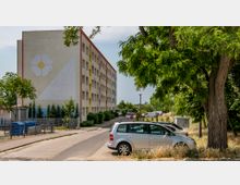 Ein vierstöckiges Wohngebäude mit heller Fassade, auf der eine große gemalte Blume und ein Dreieck zu sehen sind, flankiert von einem Gehweg, geparkten Autos und dichter Vegetation mit großen Bäumen am rechten Bildrand. Im Vordergrund befindet sich eine kleine Abfallstation mit Mülltonnen.