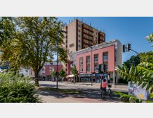 Straßenszene in einer Stadt mit einem mehrstöckigen Wohn- und Geschäftshaus in Gelb- und Rosatönen, darunter Geschäfte und Balkone. Im Vordergrund stehen Bäume, eine Ampel und ein Fahrradfahrer auf einem Radweg.