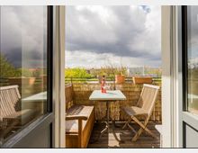 Ein kleiner Balkon mit Holzboden, ausgestattet mit einer Holzbank, einem Tisch und einem Stuhl. Auf dem Tisch stehen eine Flasche Wein und ein Glas, im Hintergrund sind Pflanztöpfe auf der Balkonbrüstung sowie eine Aussicht auf ein Wohnviertel mit Bäumen und Häusern sichtbar.