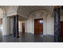 Eleganter Innenraum mit einer hohen gewölbten Decke, verziert mit Stuckarbeiten und floralen Ornamenten. Der Raum enthält dunkle Marmorsäulen mit verzierten Kapitellen, glänzende Holztüren und glatten Steinboden.